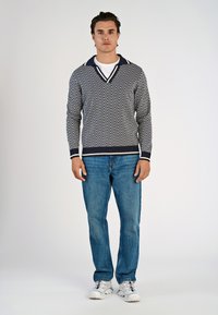 Maglione a righe zigzag blu navy e bianco con un ampio colletto, abbinato a jeans azzurri e sneakers bianche. Design semplice ma definito.