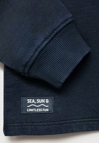 Námornícky modrý sveter s rebrovanými manžetami, ktorý má našitú etiketu s nápisom "SEA, SUN & LIMITLESS FUN" v bielom texte na tmavom pozadí.