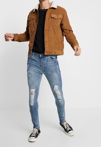 Brązowa kurtka ze sztruksu z kołnierzem z baranka, czarna koszula, jasnoniebieskie obcierające rurki oraz czarne trampki.