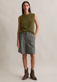Olijfgroen mouwloos topje gecombineerd met grijze geruite shorts; bruine enkellaarsjes; gladde textuur; relaxte pasvorm; knoopdetail bij de taille.