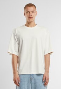 Fehér unisex póló rövid ujjakkal, puha anyagból készült, laza illeszkedésű és kerek nyakkivágású, világoskék farmernadrággal párosítva.