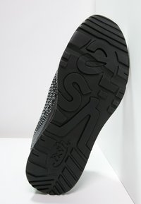 Černá podrážka tenisky z texturovaného gumového materiálu s výrazným vzorem výstupků a znakovým ražením. Tkaný svršek má skvrnitý design.