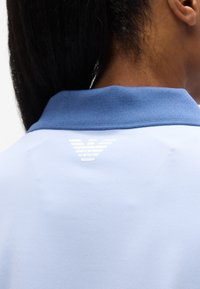 Γυναίκα με σκουρόχρωμα μαλλιά που φοράει ανοιχτό μπλε πουκάμισο πόλο με σκούρο μπλε γιακά και λευκό λογότυπο αετού στη ραχοκοκαλιά του λαιμού.