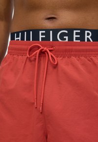 Κόκκινα μαγιό με κορδόνι στη μέση, με μπλε σκούρο λάστιχο μέσης που φέρει λευκά γράμματα "HILFIGER". Το ύφασμα έχει λεία υφή.