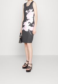 Šedé šaty bez rukávů s růžovým květinovým potiskem, vyrobené z hladkého materiálu. V páru s černými sandály na platformě s otevřeným vnitřkem a malou průhlednou taškou.