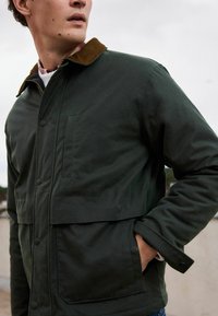 Dunkelgrüne, wetterfeste Jacke mit einem Cordkragen, Druckknopfverschlüssen, Brusttasche und elastischen Bündchen; entworfen für funktionale Schichtung.