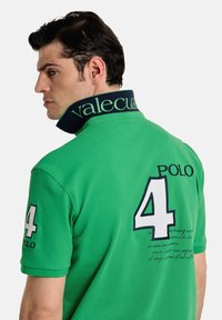 Camisa polo verde con un cuello de contraste en azul marino, bordados el '4' y 'POLO' en la espalda, con detalles en texto cursiva.