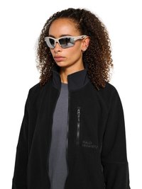 Kvinde med krøllet hår, iført grå sports-solbriller, en sort lynlåstrøje med brystlomme og en grå skjorte under.