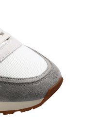 Zapatilla deportiva con parte superior de malla blanca, detalles en ante gris y suela de goma texturizada en marrón. Diseño de punta redondeada con detalles de costura.