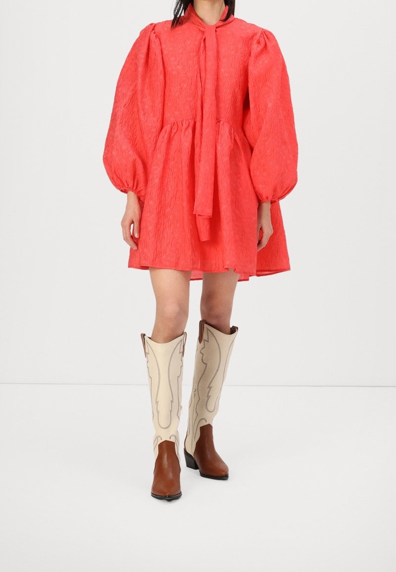 Femme portant une mini-robe rouge à manches longues bouffantes et des bottes de cowboy bicolores montantes jusqu'aux genoux, debout sur fond blanc.