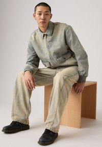 Τζιν σακάκι και παντελόνι σε ξεθωριασμένο μπλε με εμφανείς ραφές. Μαύρα παπούτσια με κίτρινες λεπτομέρειες. Το μοντέλο κάθεται σε ένα ξύλινο σκαμπό.