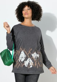Maglietta grigia a maniche lunghe con grafiche di fantasmi e foglie in marrone e bianco all'orlo, con una vestibilità rilassata e scollatura a girocollo. Borsa metallica verde.