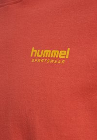 Tela roja con el logo amarillo "hummel SPORTSWEAR" impreso, destacando el nombre de la marca y el tipo de ropa.