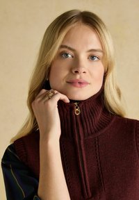 Bordeauxfarbener, gestrickter Pullover mit hohem Kragen und halbem Reißverschluss, der strukturierte vertikale Linien und goldene Reißverschluss-Beschläge aufweist.