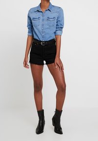 Vaaleansininen denim-paitapusero, jossa on kaksi rintataskua. Yhdistetty mustiin korkeavyötäröisiin shortseihin ja mustiin kärkipohjaisiin nilkkureihin.
