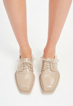 Jambes portant des chaussures à lacets beiges brillantes à bout carré, avec les lacets noués lâchement et un lacet reliant les deux chaussures.