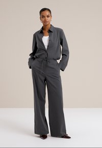 Modèle dans un ensemble gris sur mesure avec une chemise à boutons et un pantalon évasé, se tenant avec confiance devant un fond beige clair.
