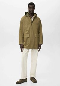 Chaqueta impermeable verde oliva con capucha, cierre frontal, dos grandes bolsillos; combinada con pantalones en tono marfil y zapatos marrones. Fondo liso.
