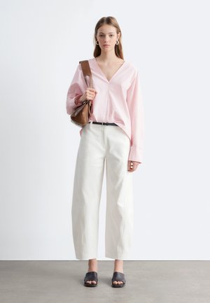 Joven mujer de pie, vestida con blusa rosa claro con escote en V, pantalones blancos de pierna ancha, zapatos negros abiertos en la punta, sosteniendo un bolso marrón al hombro sobre fondo blanco.