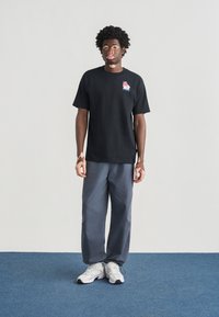 Μαύρο βαμβακερό μπλουζάκι με γραφικό σχέδιο στο στήθος, σε συνδυασμό με ανοιχτό γκρι φαρδιά παντελόνια και λευκά αθλητικά παπούτσια πάνω σε μπλε χαλί.