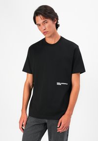 Camiseta de algodón negra con cuello redondo, mangas cortas y un estampado de logo blanco en el lado izquierdo del pecho. Ajuste simple y relajado.