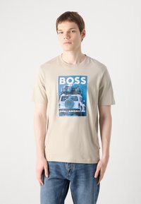Tricou din bumbac bej cu o imprimare grafică albastră a unui vehicul, având textul "BOSS" și "PAN-AMERICAN". Mâneci scurte, design cu guler rotund.