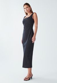 Žena u crnoj bezrukavnoj midi haljini s bijelim točkicama i crnim sandalama na petu, stoji na jednobojnoj bijeloj pozadini.