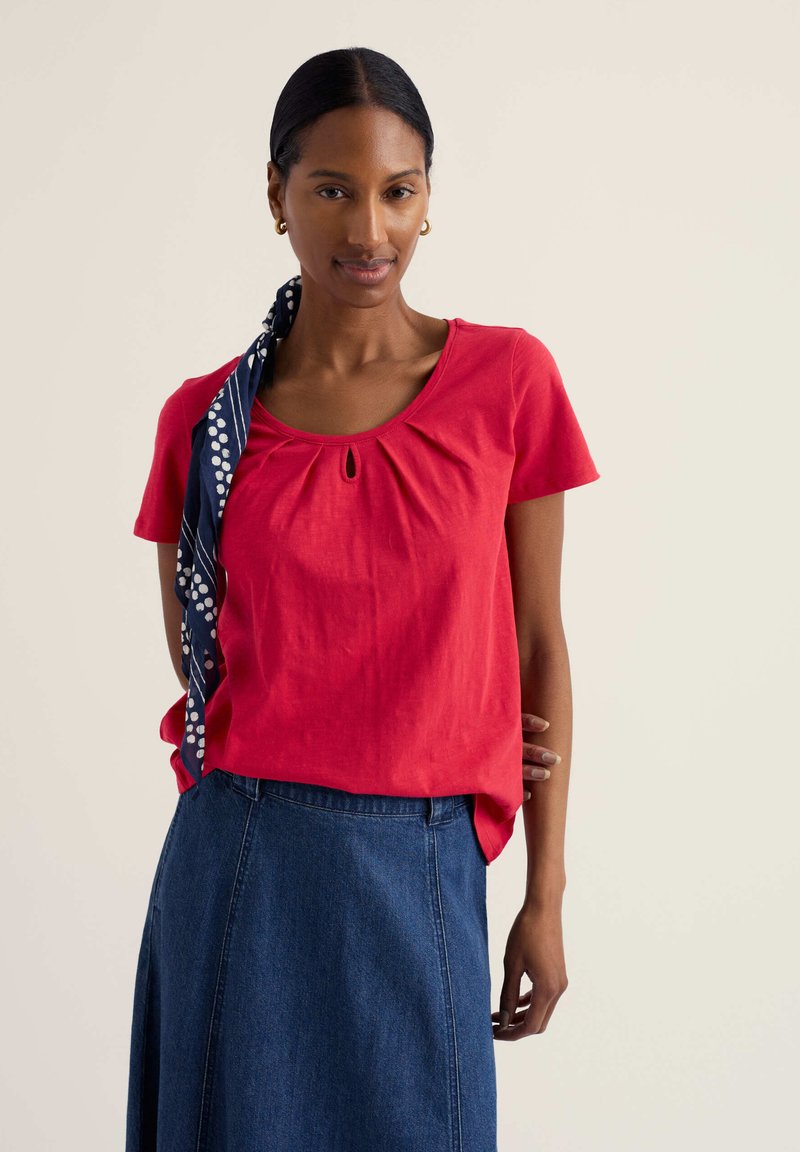 Femme aux cheveux foncés attachés, portant un haut rouge à manches courtes, une jupe en jean bleue, des boucles d'oreilles en or en forme de créole et un foulard marine à pois.