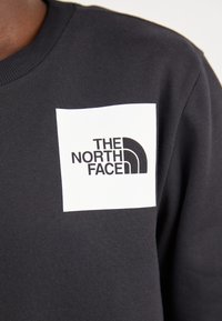Svart hettegenser med en rektangulær hvit lapp med "THE NORTH FACE"-logo i svart tekst og en buet grafikk.