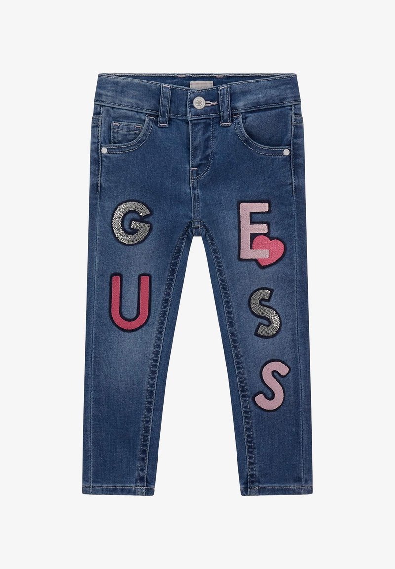Kék farmernadrág színes hímzett betűkkel és szimbólumokkal: "G", "U", "E", "S", szívmotívummal és texturált részletekkel.