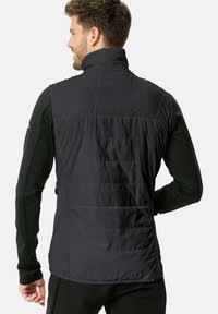 Gilet isolato nero con alto colletto, texture trapuntata e pannelli laterali. Indossato sopra un top a maniche lunghe, che mette in mostra un design elegante e aderente.