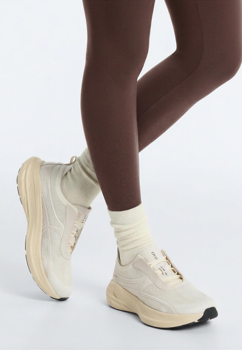 Personne portant des baskets montantes beiges à semelles épaisses et des chaussettes crème clair, debout jambes croisées devant un fond uni.