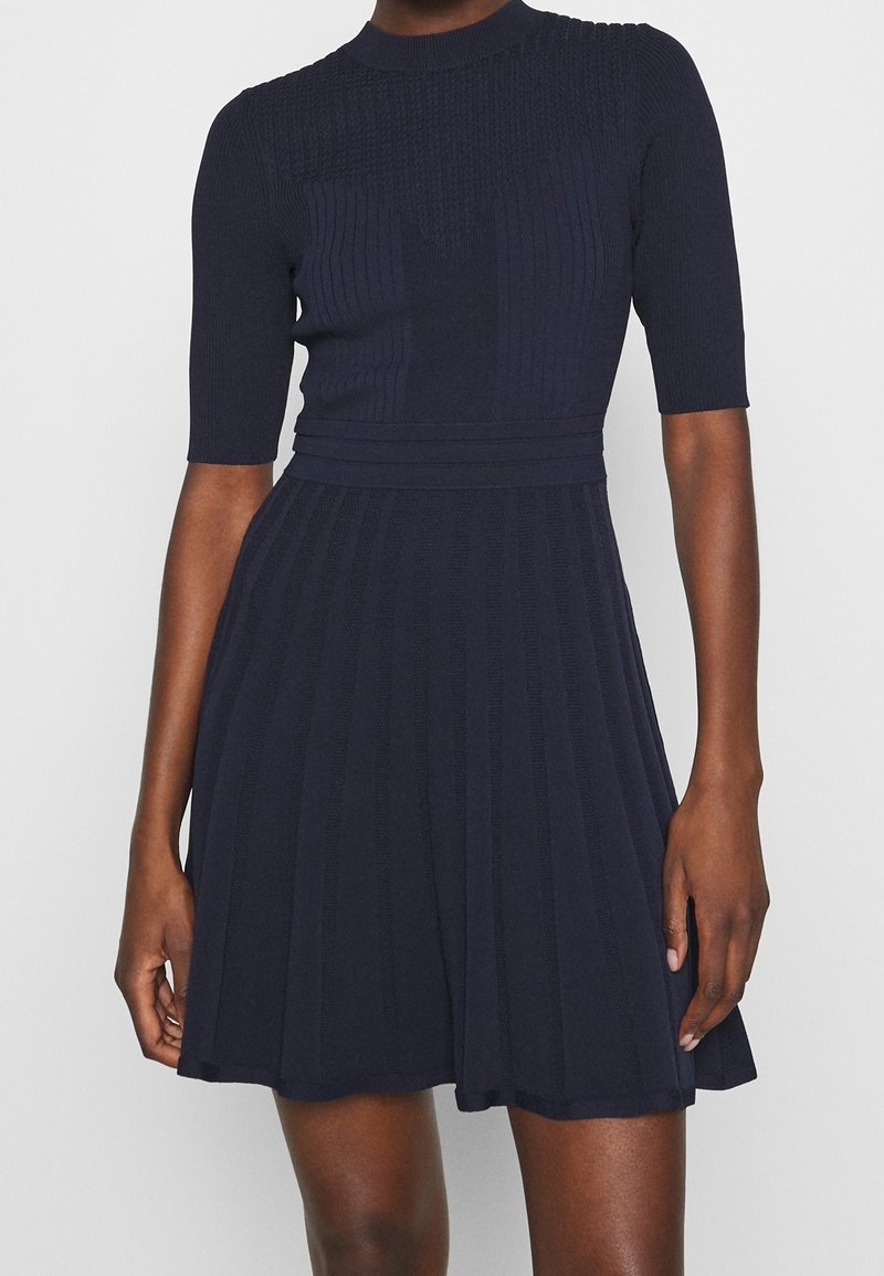 Femme portant une robe en maille bleu marine à manches courtes, taille cintrée et jupe évasée sur un fond uni gris clair.