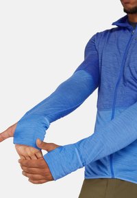 Giacca atletica blu a maniche lunghe con zip, caratterizzata da tonalità contrastanti e aperture per il pollice, realizzata in un tessuto leggero e testurizzato.