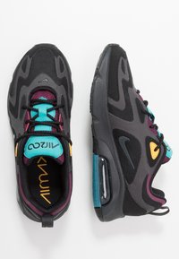 Pari mustia ja harmaita lenkkareita, joissa on teal-, violetin ja keltaisia yksityiskohtia, mustat kengännauhat ja näkyvä ilmapatja valkoisella taustalla.