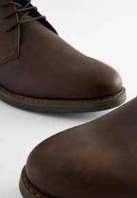 Zapatos de cuero marrón oscuro con punta redonda, textura suave y detalles de costura a lo largo de la suela, con cordones y un perfil bajo.