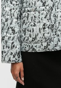 Απαλή μπουφάν σε ανοιχτό γκρι χρώμα με μαύρα αφηρημένα μοτίβα, διαθέτοντας μία τσέπη με κουμπώνει και πλικέ υφή, παρουσιασμένο με το χέρι πάνω στο μανικί.