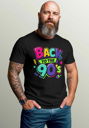 Bald man with gray beard, tattoos, and watch wears black shirt reading "Back to the 90's" in colorful bold letters, hands in jeans pockets.