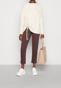 Pull en maille couleur crème avec détail froncé sur le côté, associé à un pantalon court marron foncé, des baskets blanches et un sac fourre-tout beige.