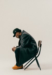 Un homme en manteau sombre, portant une casquette noire et des bottes beiges, est assis penché en avant, les mains jointes, sur une chaise en bois devant un arrière-plan uni.