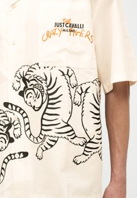 Μπεζ πουκάμισο με κοντά μανίκια, διακοσμημένο με μαύρες απεικονίσεις τίγρεων και πορτοκαλί-μαύρο κείμενο στην τσέπη που γράφει «The Just Cavalli Milano Crazy Tigers».