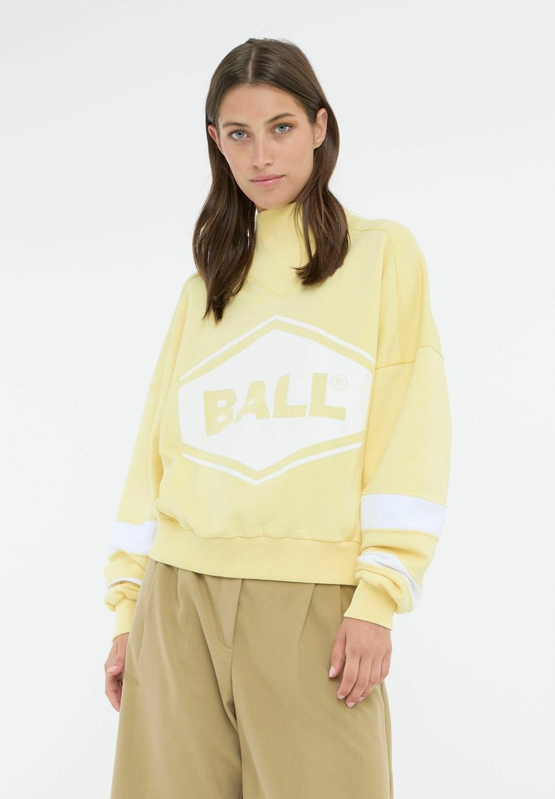 Gul oversized sweatshirt med et hvidt "BALL" logo, ribbet krave og hvide stribede detaljer ved manchetterne. Parret med løse beige bukser.