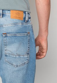 Jasnoniebieskie dżinsy z denimu z prostokątną skórzaną naszywką na tylnej części pasa, ozdobione przeszyciami i pojedynczą tylną kieszenią.