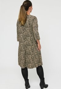 Vrouw met lang bruin haar in een staart, gekleed in een geblokte jurk tot de knie, zwarte maillot en zwarte enkellaarzen, staande met de rug naar de kijker gekeerd.