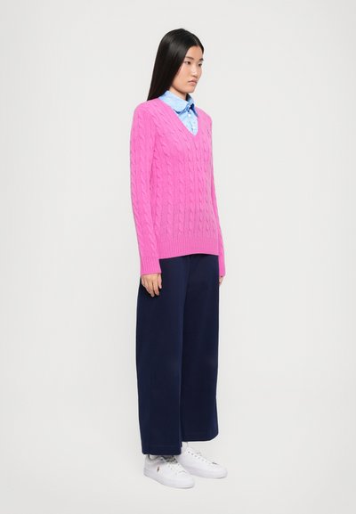 Roza pletena jopica z V-izrezom in pletenim vzorcem, v kombinaciji s širokimi modrimi hlačami in belimi supergami. Pod njo je svetlo modra srajca s kratkimi rokavi.