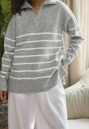 Grijze gestreepte trui met een halve ritskraag, gemaakt van zacht materiaal, met witte horizontale strepen en een losse pasvorm. Gecombineerd met witte broek.