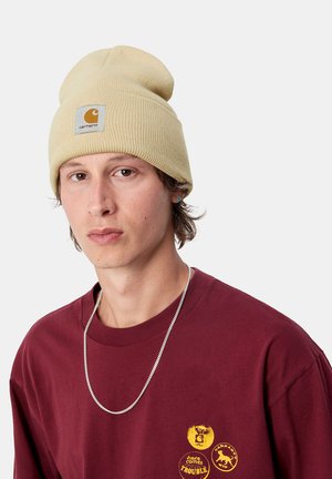 Berretto di maglia beige con risvolto e patch con logo, indossato con una maglietta bordeaux con stampe grafiche gialle e una collana in argento.
