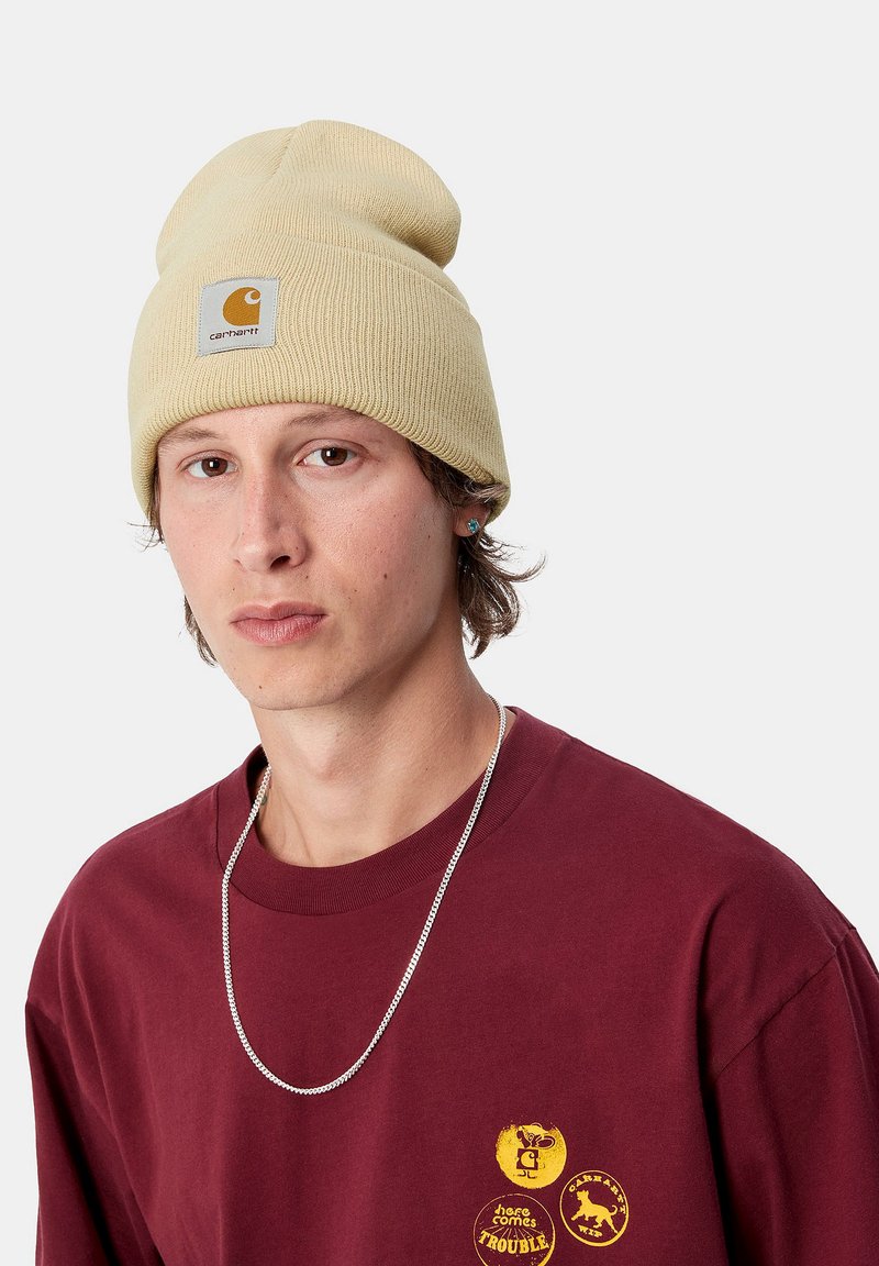 Gorro de punto beige con dobladillo y parche de logo, combinado con una camiseta marrón burdeos con estampados gráficos amarillos y una cadena de plata.