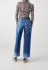 Osoba v modrých vysokých riflích s manžetami, dlouhém tričku se zebřími pruhy a teniskách se zebřími vzory, stojící na neutrálním světlejším pozadí.