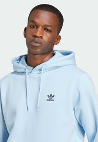 Lichtblauwe katoenen hoodie met een voorzak, een capuchon met trekkoord en een donkerblauw Adidas-logo op de linkerborst. Glad textuur.